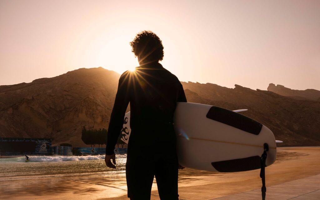 A surfer holding a board faces the wave pool at Al Ain Adventure, with desert mountains behind at sunset