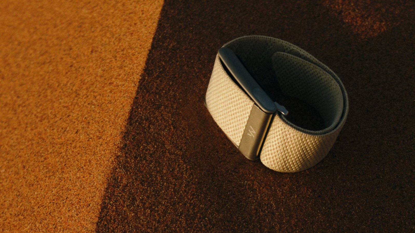A gold-toned Whoop band resting on a textured tan surface in warm light